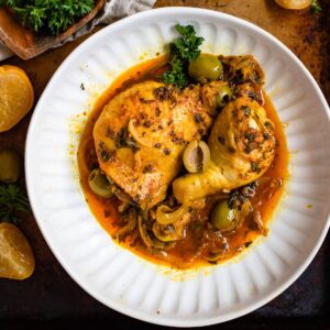 Moroccan chicken tagine served on a plate with vegetables using chicken thighs in a flavorful broth with halved green olives garnished over it.