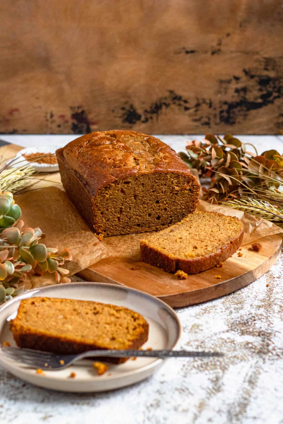 Pumpkin bread on a wooden cutting board with a slice cut away in front of it and anotherslice on a small plate to the side. 