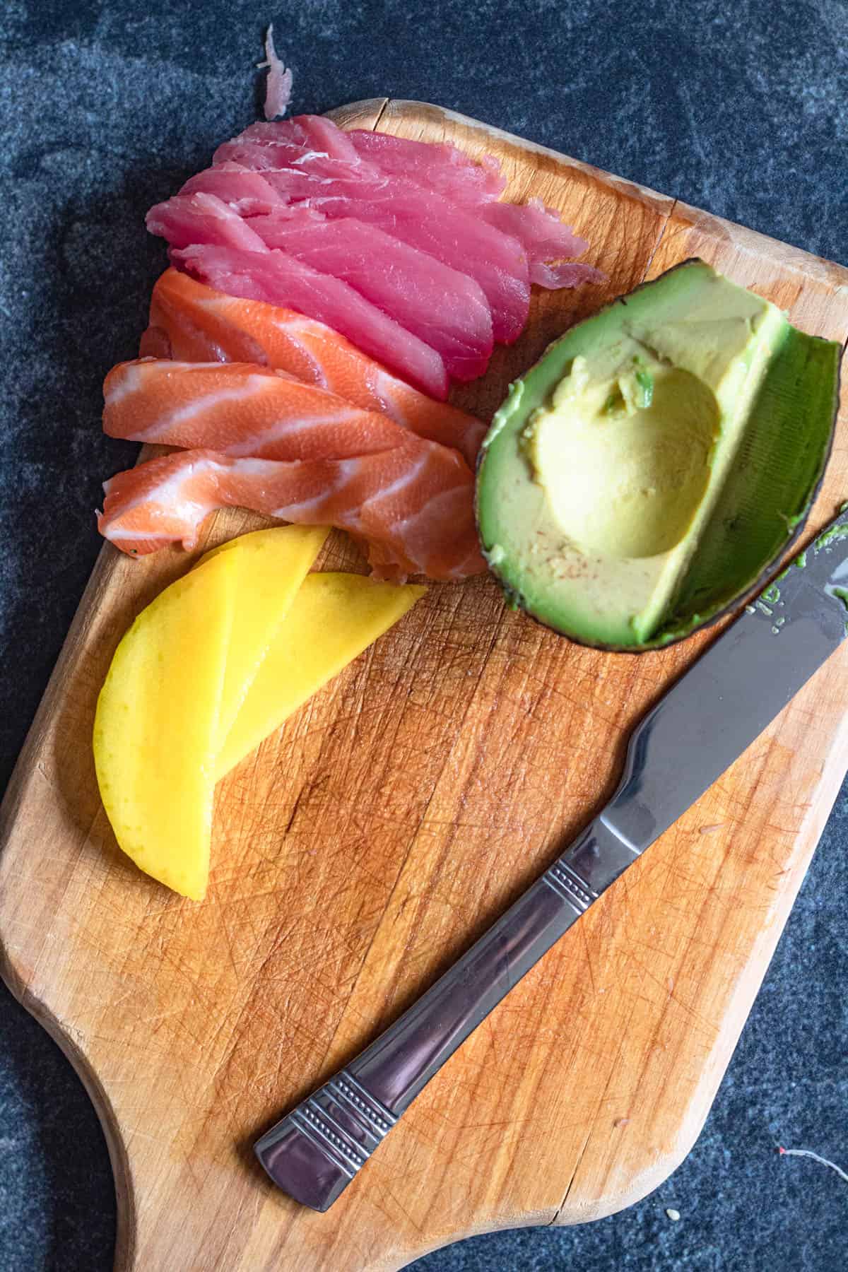 Cutting board showing thinly sliced tuna, salmon with mango slices and half an avocado. 