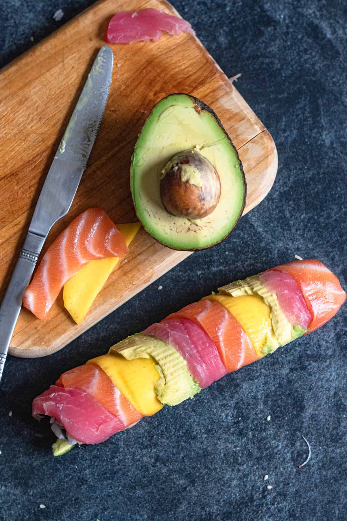 Sushi rice rainbow roll covered with thinly sliced tuna, salmon, mango and avocado sitting next to a cutting board with leftover ingredients. 