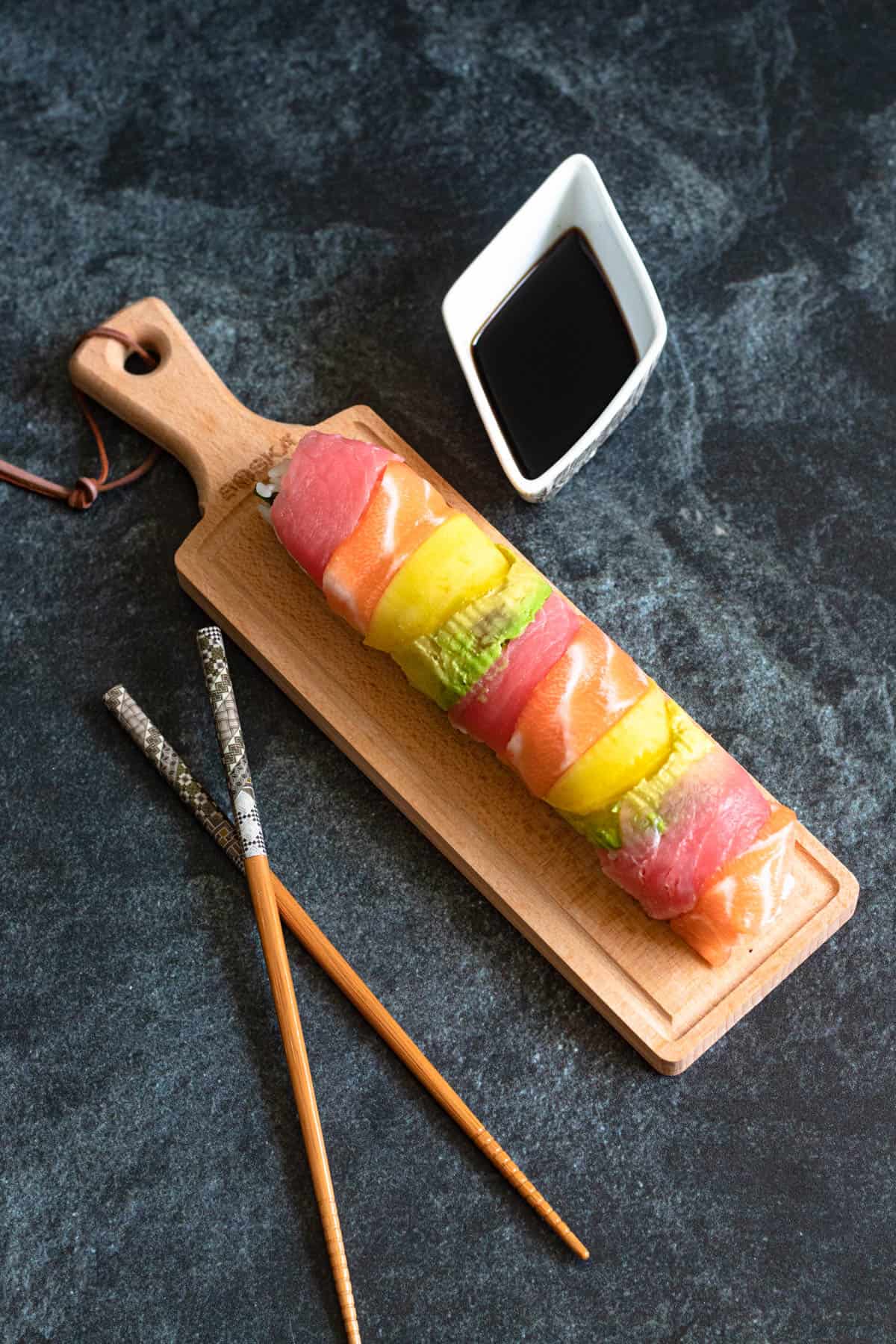 A small bowl of dipping sauce next to the rainbow roll sushi laying on a serving board with a pair of chopsticks laying next to it. 