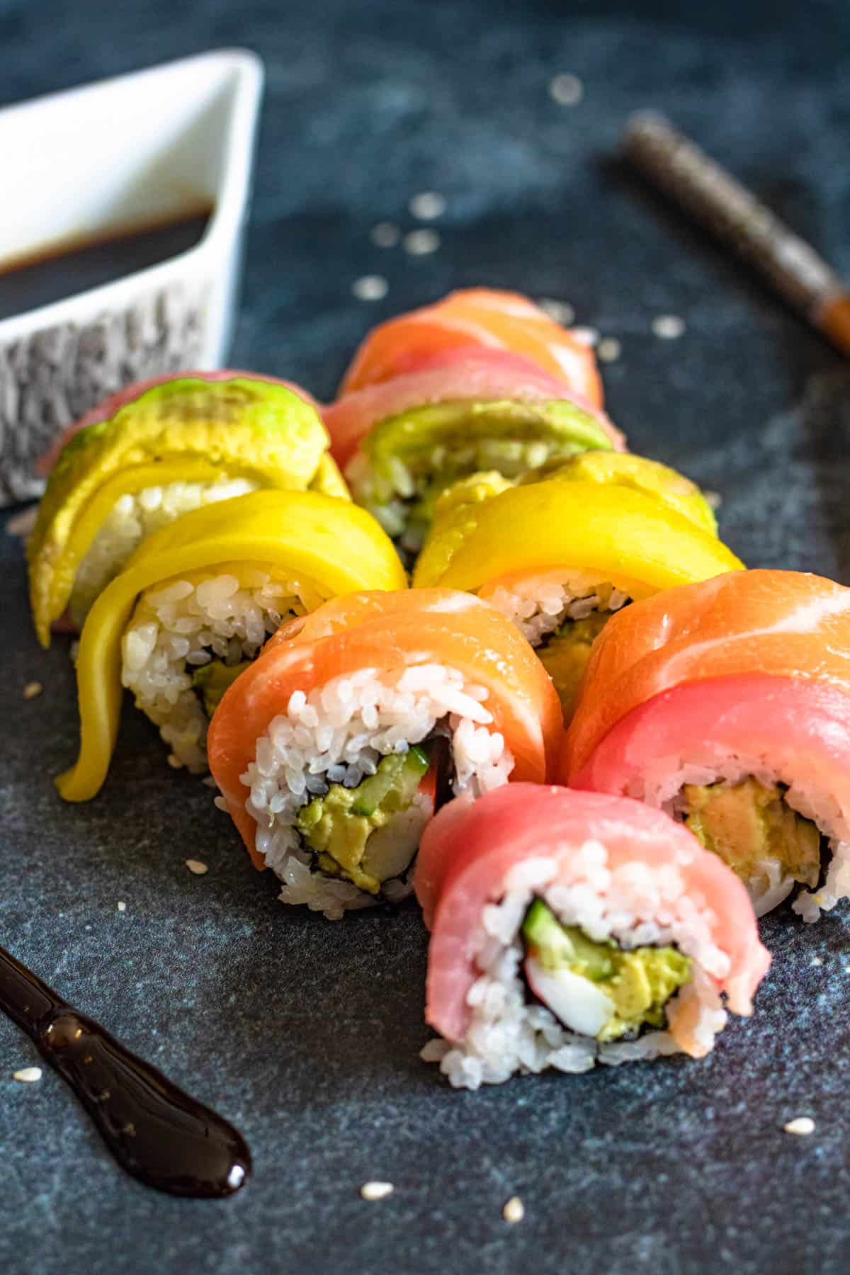 Pieces of Rainbow roll sushi sitting on a serving board, with chopsticks and soy sauce behind them. 