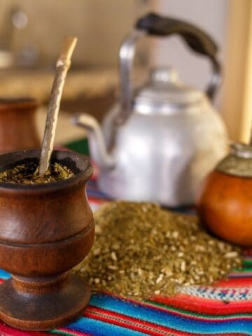 A small wooden mortar with yerba mate being crushed and fresh tea leaves spilled over a colorful blanket that has a tea pot laying behind it.