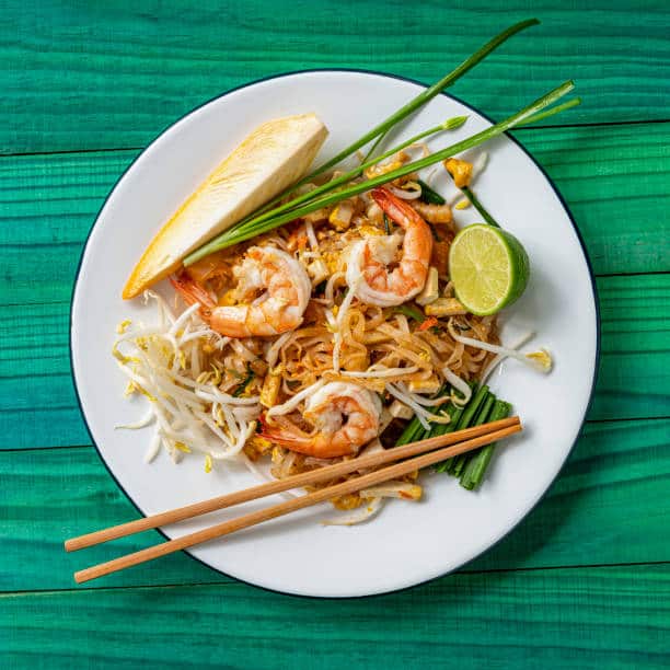 Chopsticks laying on the side of plate with prawn pad thai with a half lime garnished on the side. 