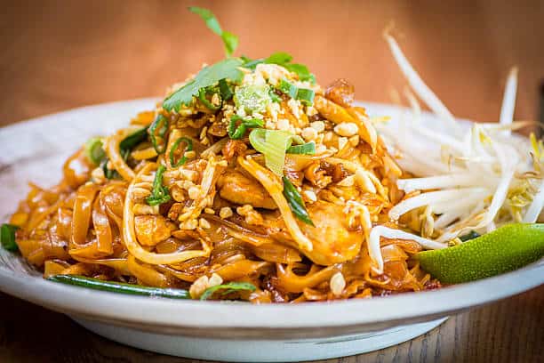 Pad thai on a plate with chopped peanuts and bean sprouts with cilantro chopped on the side. 