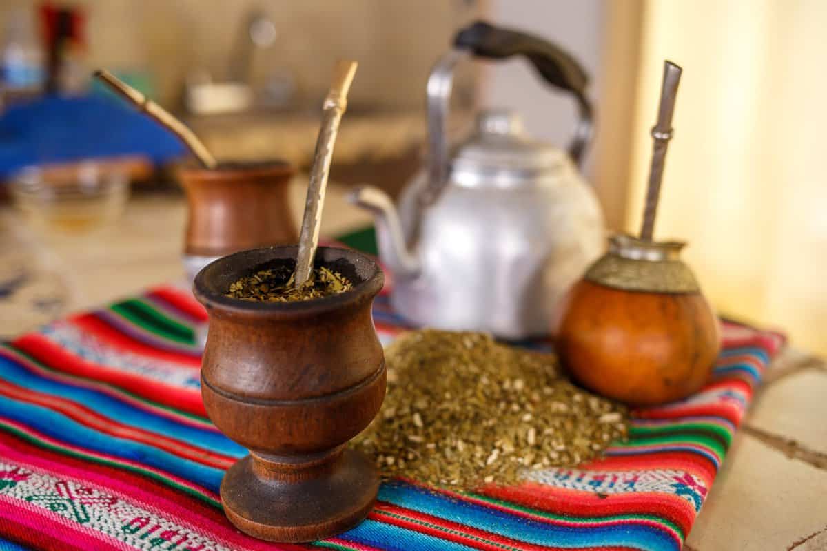 Yerba mate served in gourd and a metal straw coming out of it, with the dry leaves laying on a colorful south american blanket with the hot water in a tea pot next to them. 