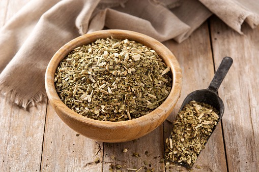 Yerba mate leaves in a small bowl with a scoop filled with the leaves laying next to it, some spilling out. 
