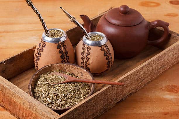 Dry yerba mate leaves in a small bowl with two gourds with metal straws inserted into the mate next to a tea pot on a serving tray. 