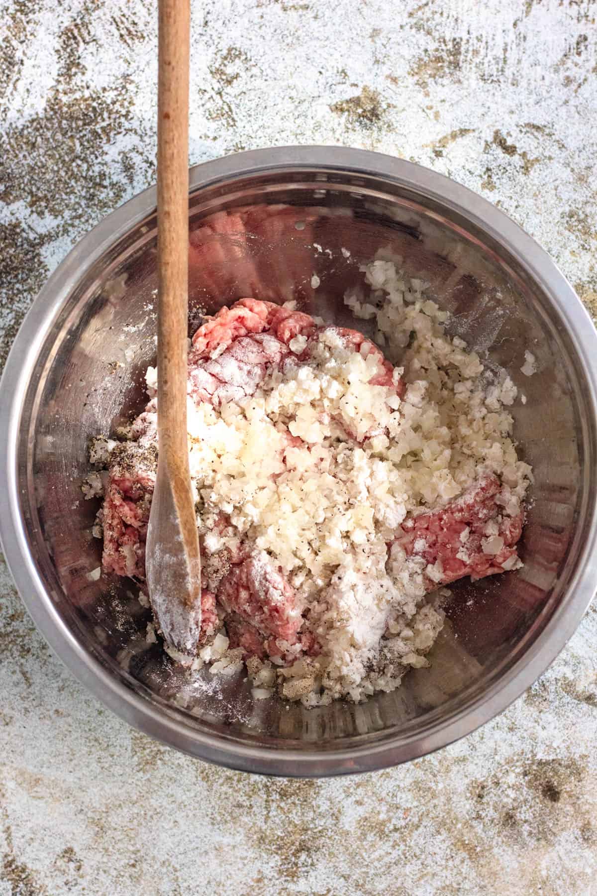 Seasonings and ground pork added to a mixing bowl with a wooden spoon resting in it. 