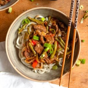 Flavorful stirfry topping homemade noodles with a variety of veggies, garnished with thinly sliced green onions and chopsticks resting on the side of the bowl.