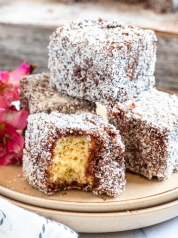 A pile of lamingtons with a bite being taken out of one, with flowers in the background.