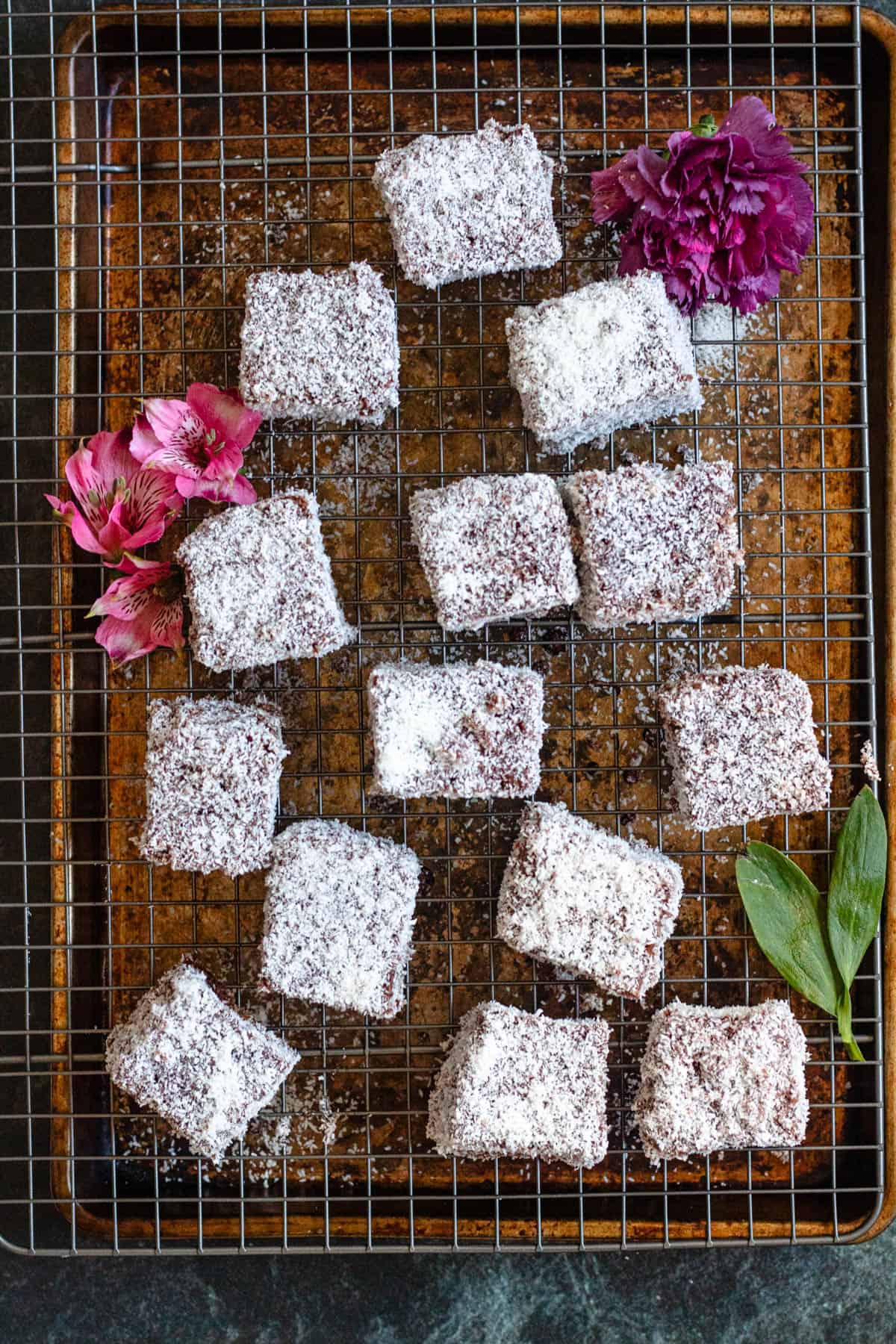 Lamingtons on a cooling rack with pink flowers garnished alongside them. 