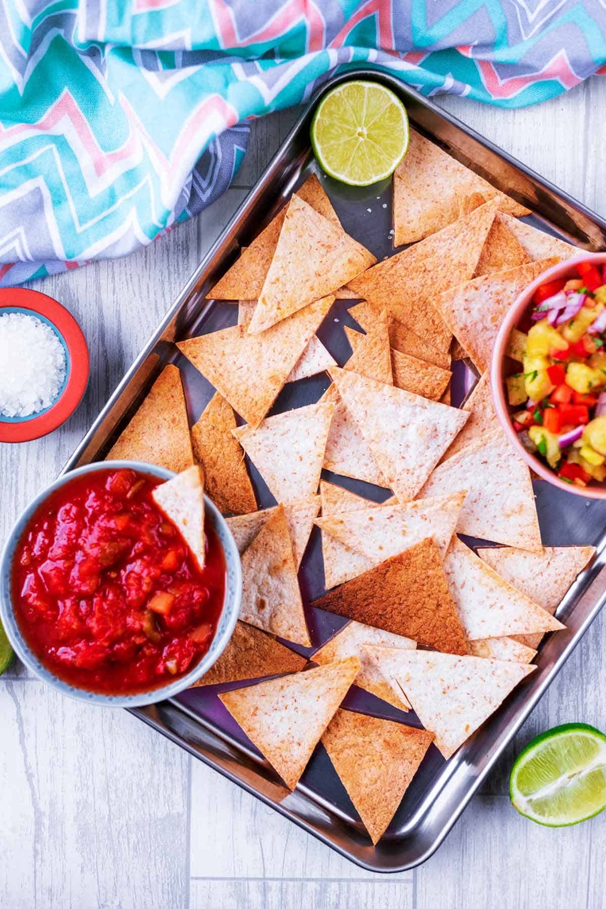 Baked tortilla chips on a baking sheet cut into triangles and a bowl of pico de gallo and one with salsa. 