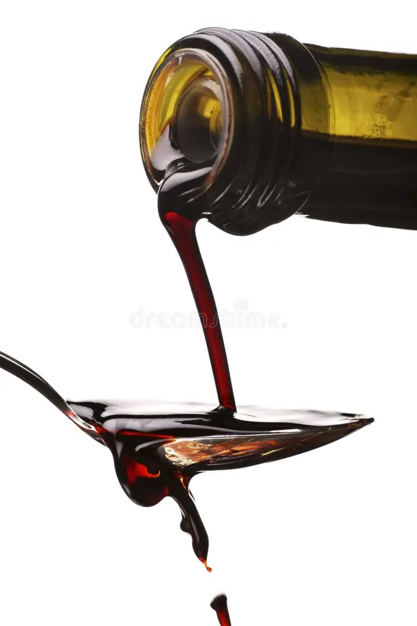 Dark brown balsamic vinegar pouring out of a glass bottle onto a spoon and spilling down off the spoon. 