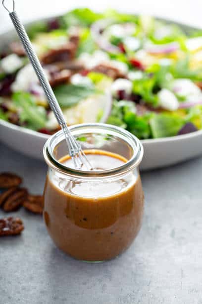 Small whisk resting in a small glass jar of balsamic vinaigrette next to a green salad in a bowl. 