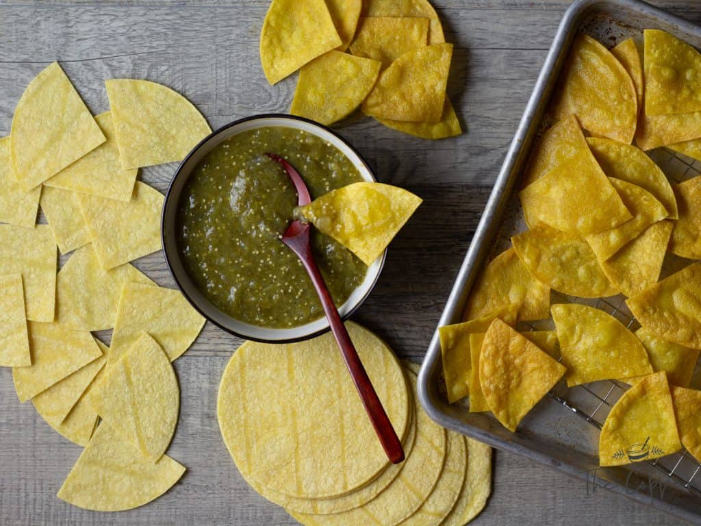 Sheet pan salsa verde in a small bowl with a spoon in it, served with corn tortilla chips baked and whole corn tortillas around the salsa. 