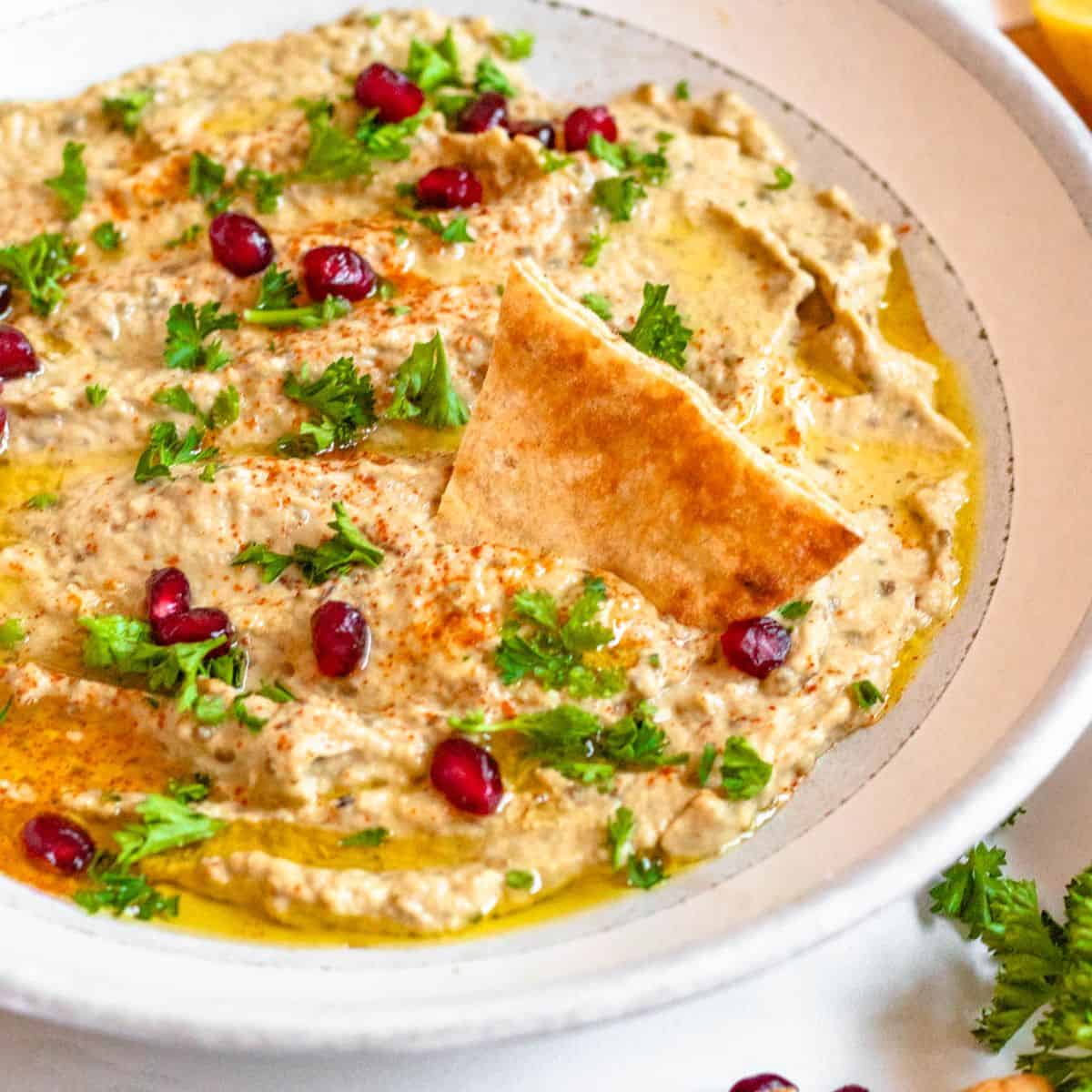 Pomegranate seeds topping baba ganoush with slice of pita bread dipping into it.