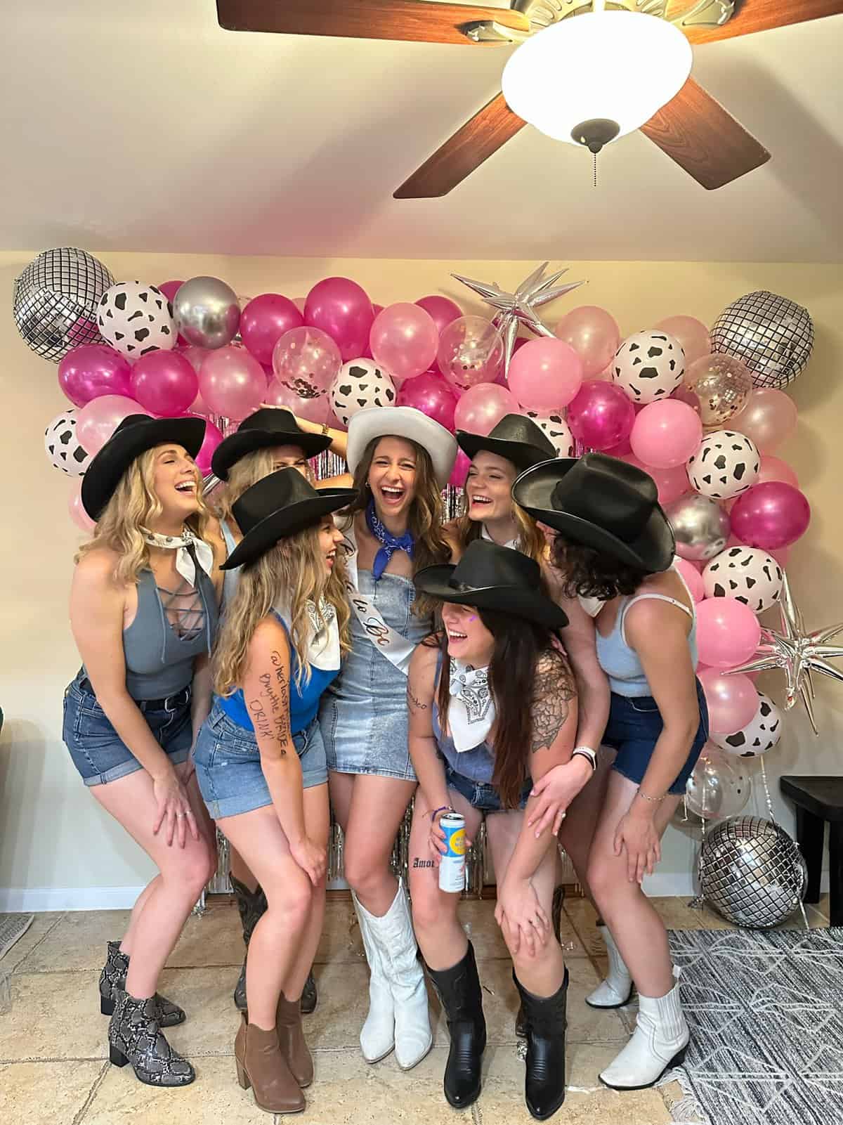 A group of girls laughing in front of a balloon arch, wearing cowboy boots. 
