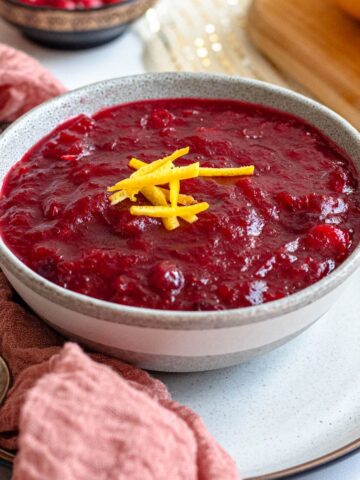 Close up of cranberry sauce topped with lemon zest and sitting next to a pink linen and a serving spoon.