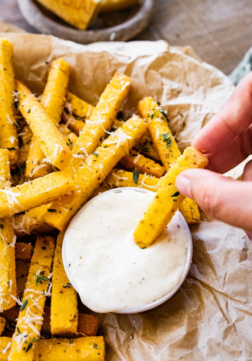 Hand picking up a crispy baked polenta fry from a basket  and into a bowl of dipping sauce. 