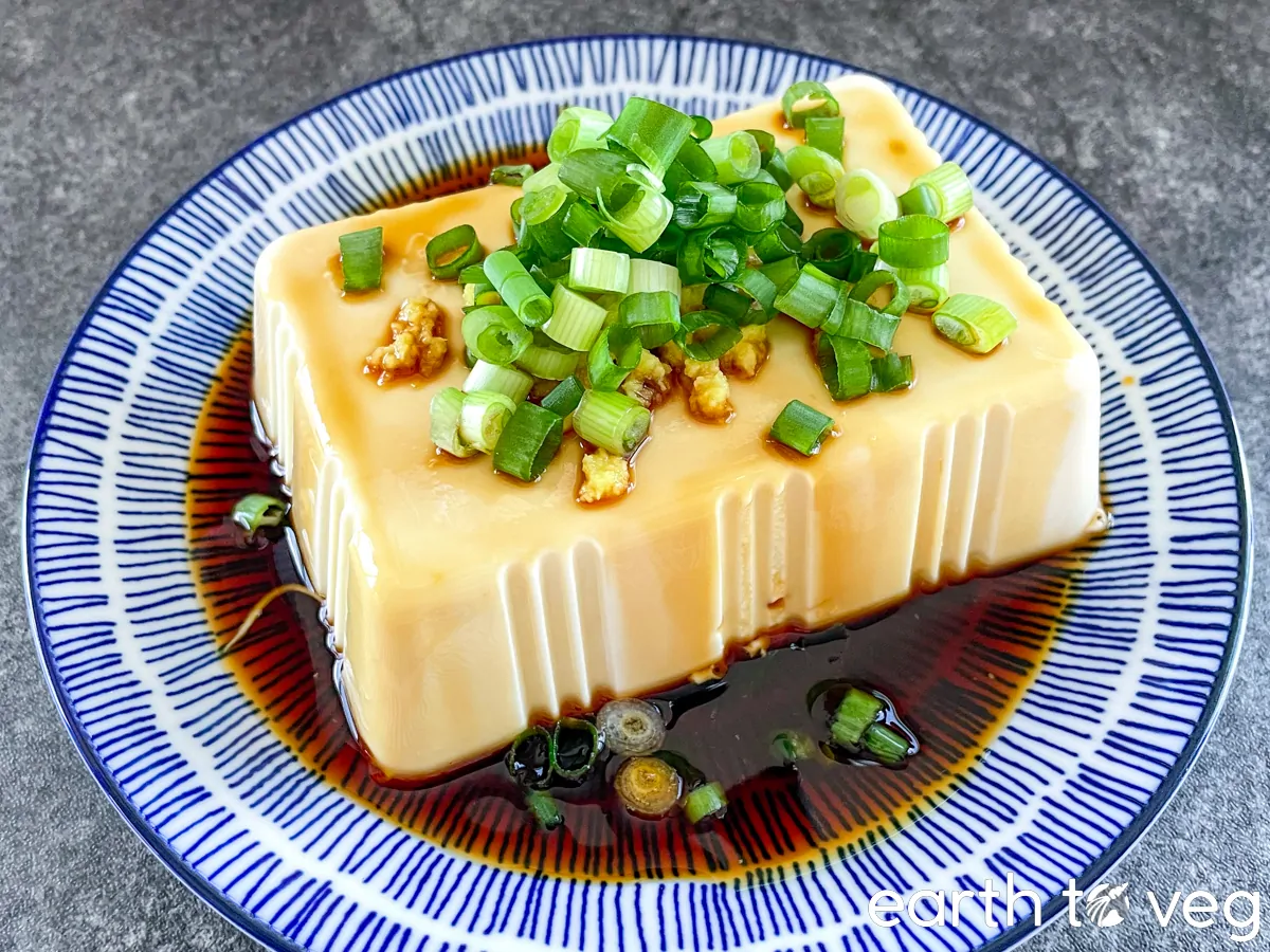 Hiyayakko on a plate topped with sliced green onions and soy sauce pooled around the bottom of the tofu. 