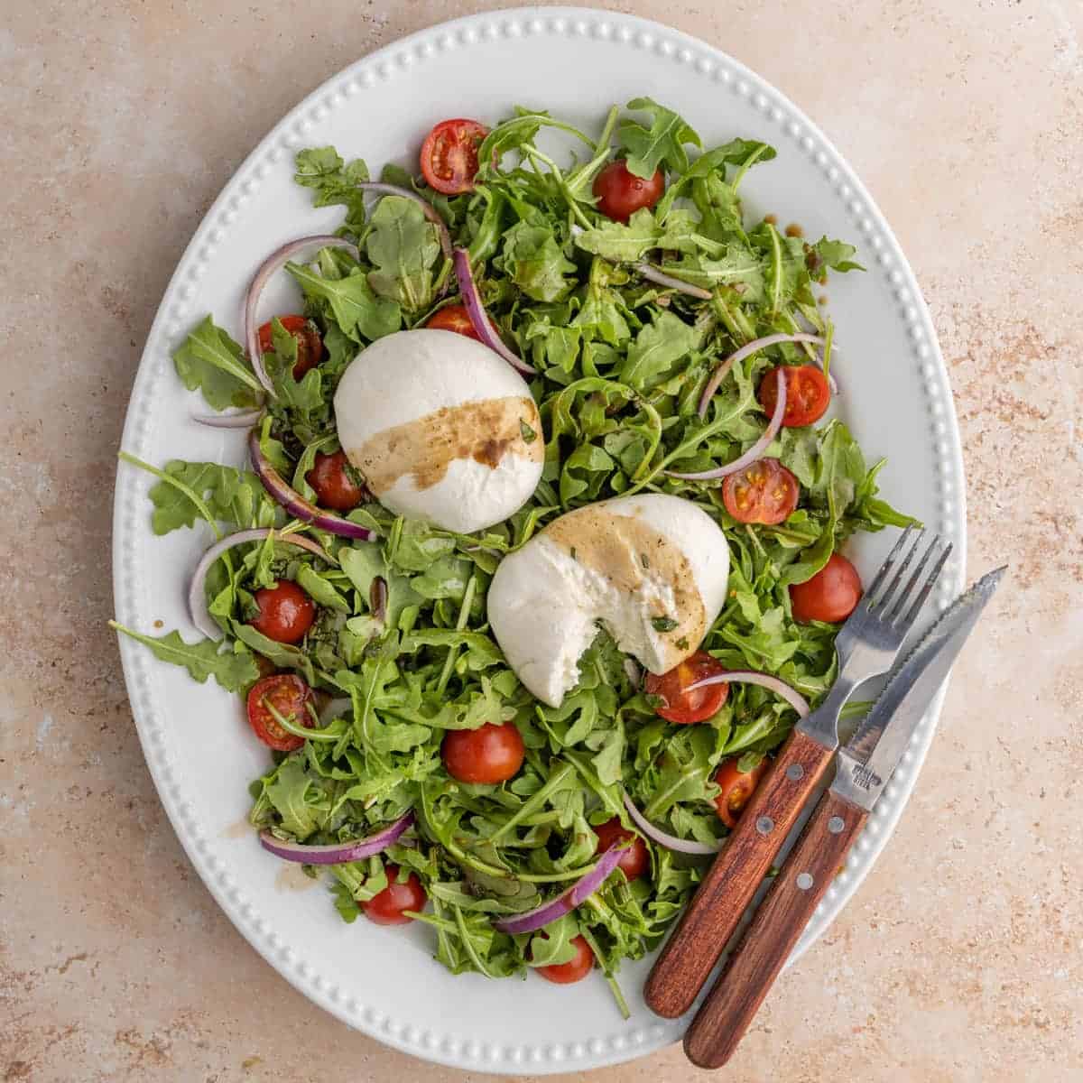 Large serving platter of arugula burrata salad with halved cherry tomatoes and red onion slices, drizzled with basil balsamic. 