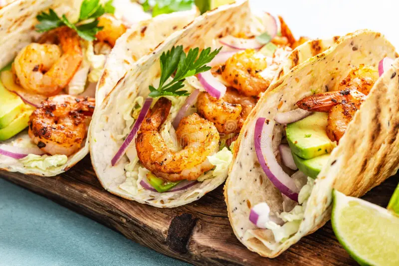 Three shrimp tacos served in grilled flour tortillas with lime wedges garnished on the side. 
