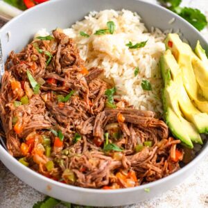 Carne Mechada, a Venezuelan shredded beef, served in a bowl next to a serving of white ricew with slices of avocado served over the top.