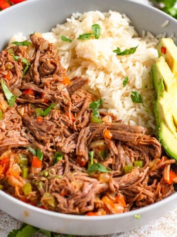 Carne Mechada, a Venezuelan shredded beef, served in a bowl next to a serving of white ricew with slices of avocado served over the top.