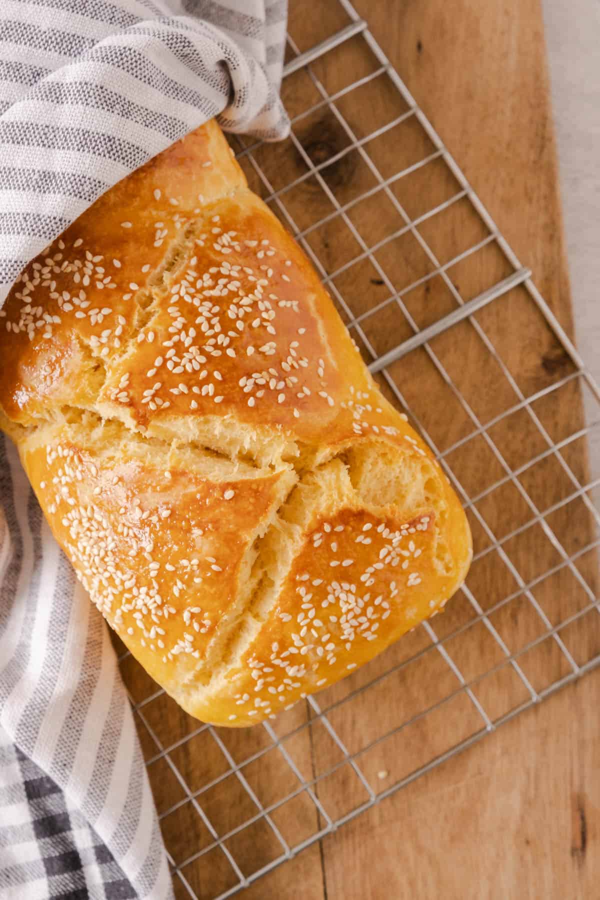 Top view of a freshly baked brioche bread loaf on a cooling rack, partially covered by a kitchen towel. 