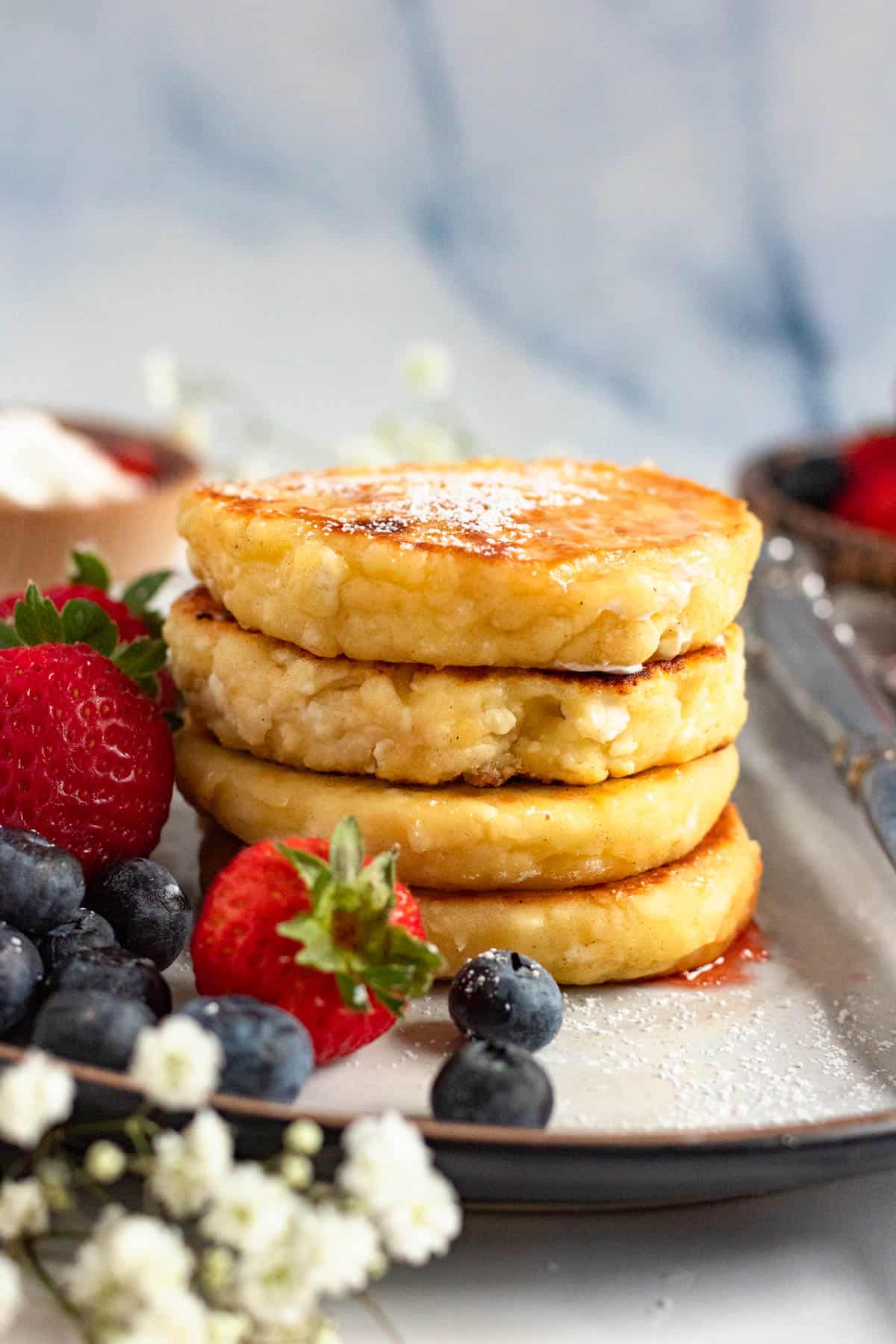 Stack of Syrniki on a plate with powdered sugar sifted over the top and fresh berries gathered around it. 