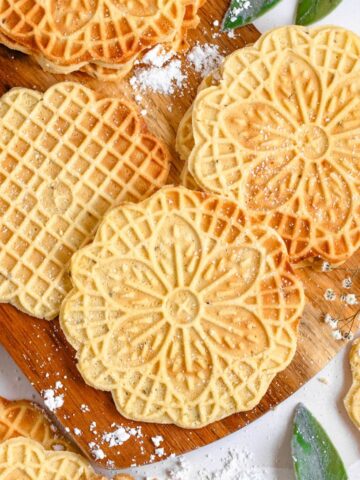 Beautiful traditional Italian Pizzelle cookies laying on a wooden serving board with powdered sugar dusted over some of them.
