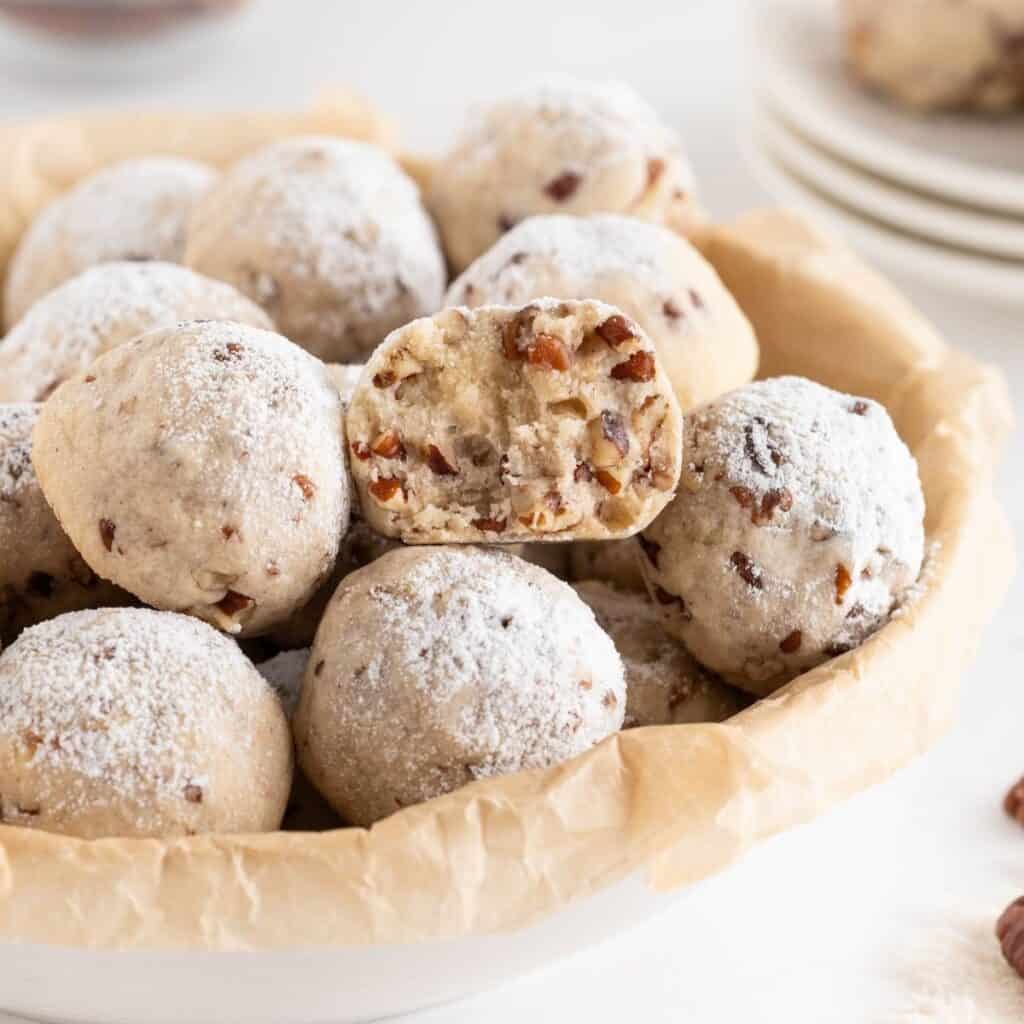 A bite taken out of a buttery pecan snowball cookie sitting on a bowlful of them.