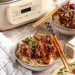 Chopsticks resting on the side of a bowl of rice topped with slow cooker Mongolian pork next to the Magnifique slow cooker.