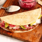 Pinterest image of a close-up shot of a Baleada ready to enjoy, and a top view shot of avocado slices over pickled onions, sour cream, cotija cheese, and refried beans on a flour tortilla.