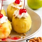 4 poached apples with whipped cream and a cherry on top served on a plate.
