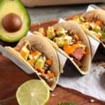 Three tacos in a taco holder with an avocado behind them.
