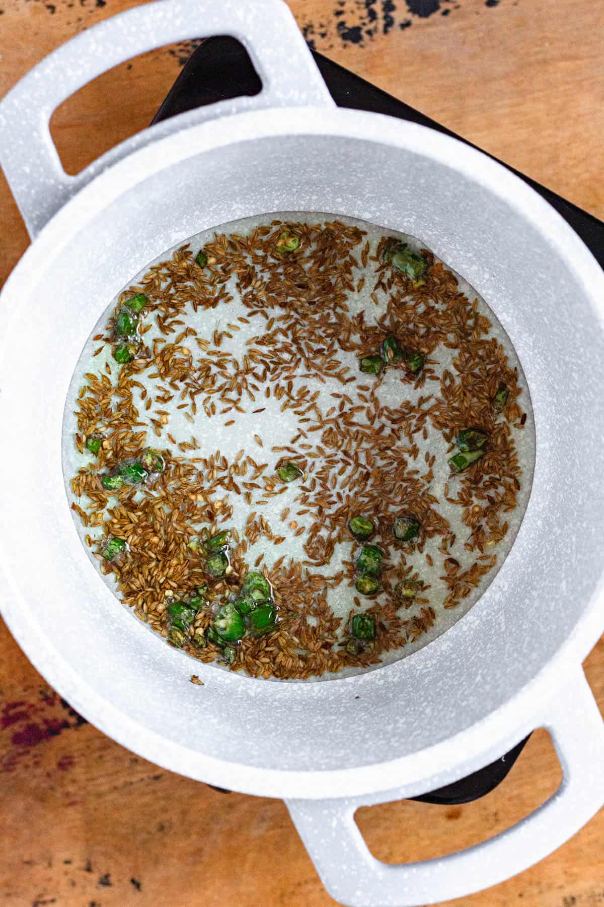 Cumin seeds and chopped green chilies sizzling in oil in a white pot.