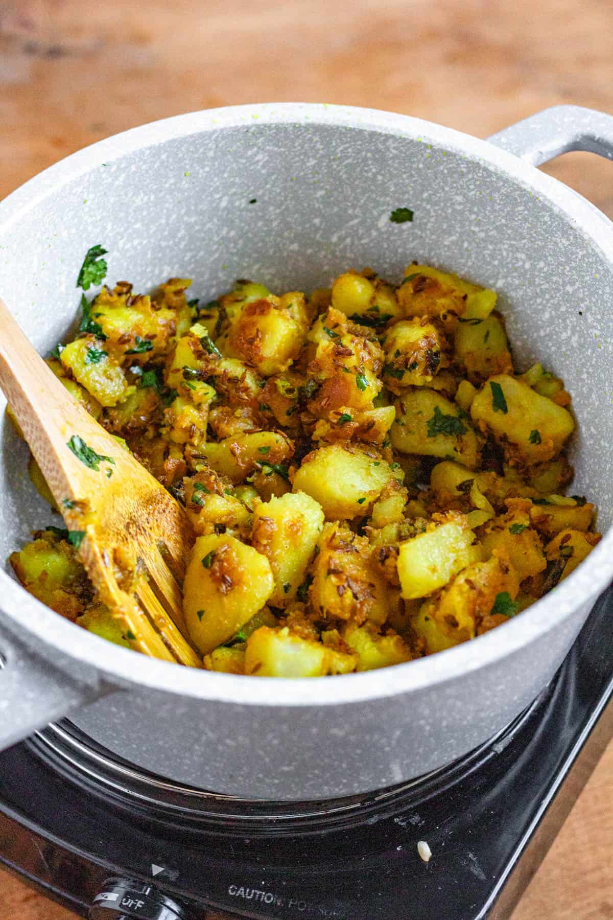 Jeera Aloo garnished with fresh coriander in a white pot, and stirred with a wooden spatula.