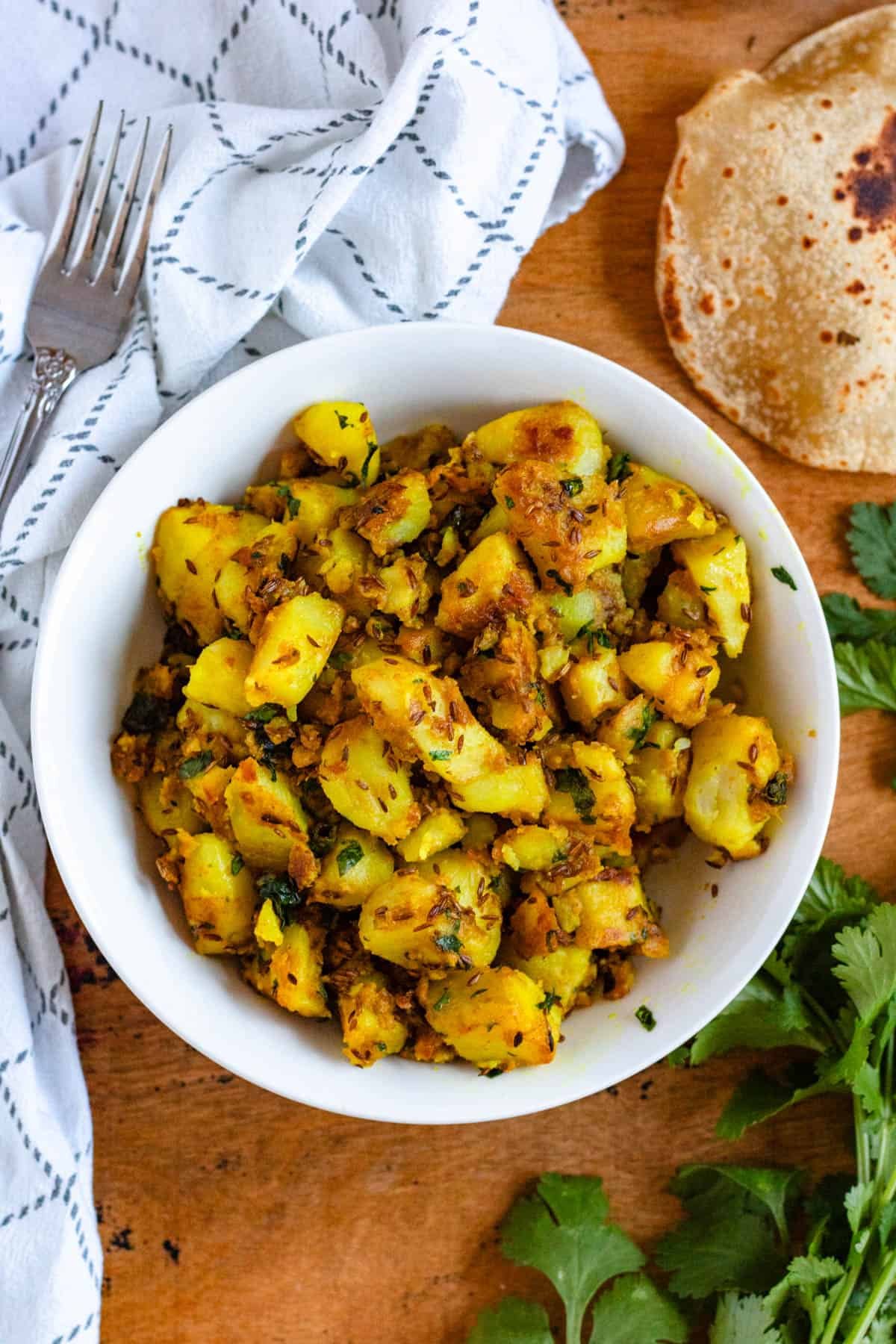 A bowl of Jeera Aloo, garnished with coriander, with roti beside it on a rustic wooden surface.
