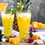 Two pineapple agua fresca drinks with mint and pineapple garnish, with whole and sliced pineapple in the background.