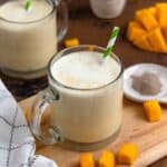 A creamy mango milk served in a clear glass mug with a green-striped straw, surrounded by fresh mango cubes and spices on a wooden board.