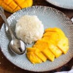 A plate of sweet mango sticky rice topped with sesame seeds, with spoon on the side.