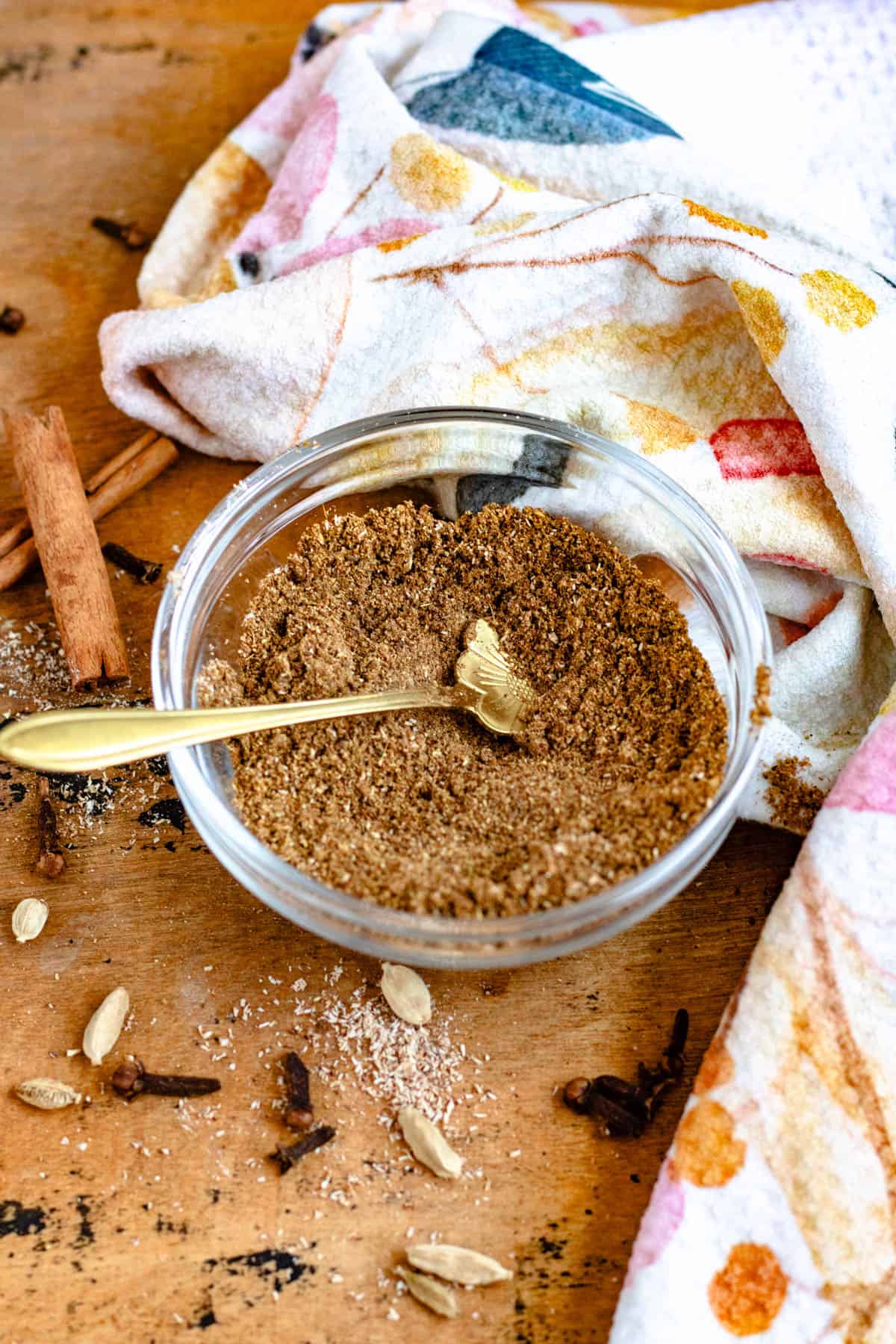 A bowl of homemade Garam Masala with a spoon, surrounded by whole cloves, cardamom pods, cinnamon sticks.