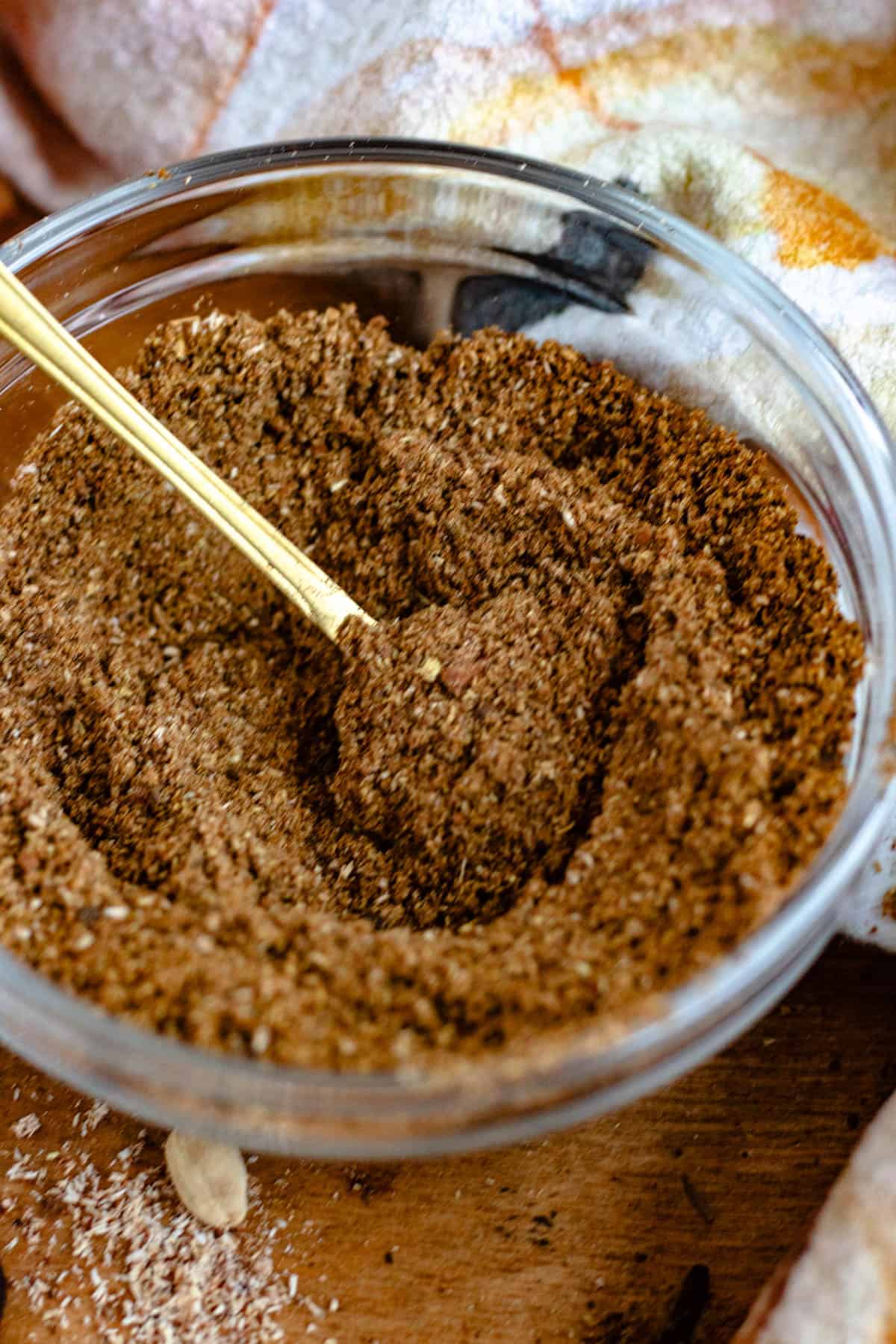 A golden spoon scoops a fragrant Garam Masala from a glass bowl, surrounded by whole spices on a wooden surface.
