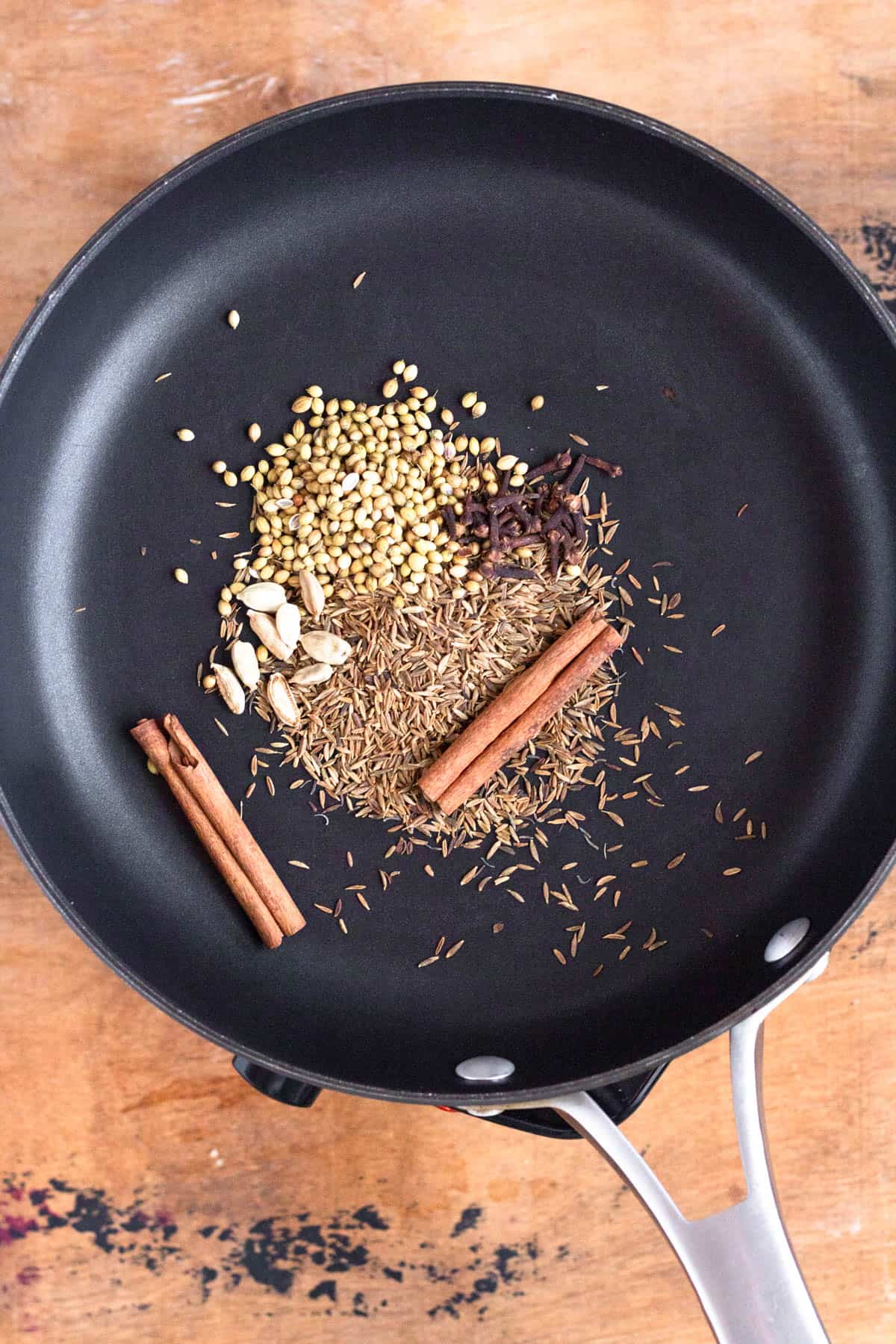 A mix of whole spices, including cinnamon sticks, cloves, cumin, cardamom, and coriander seeds, is toasting in a black skillet.