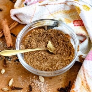 A bowl of homemade Garam Masala with a spoon, surrounded by whole cloves, cardamom pods, cinnamon sticks.