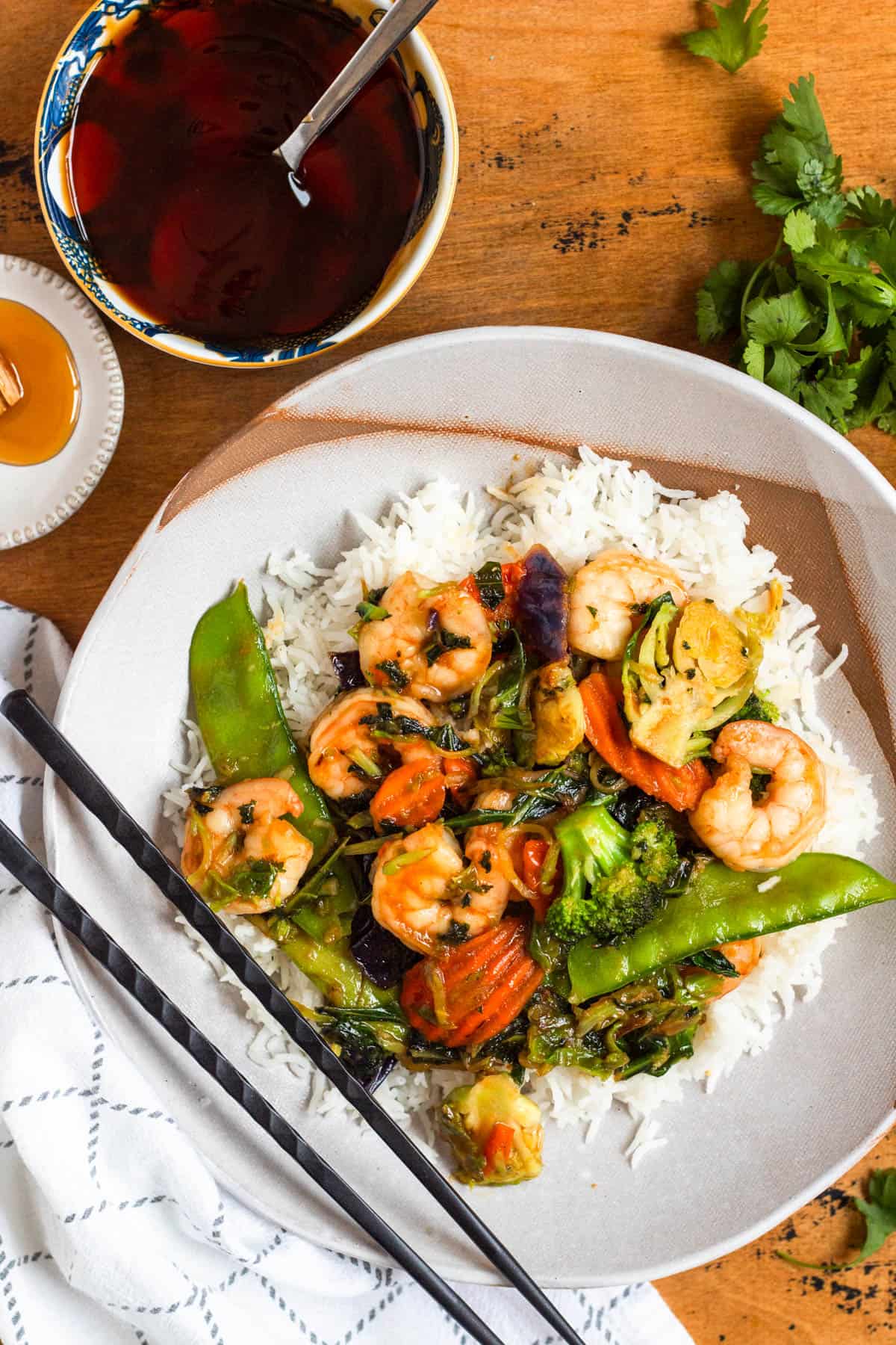 A plate of shrimp and vegetable stir-fry served over white rice with chopsticks and 3 ingredient stir fry sauce on the side.