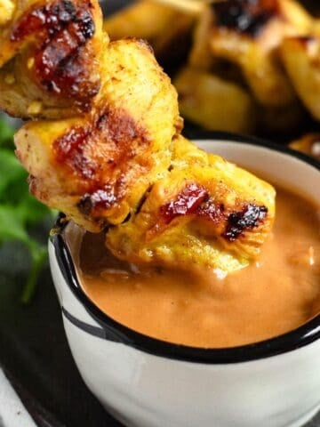 A close-up of chicken satay being dipped into a small bowl of creamy peanut sauce.