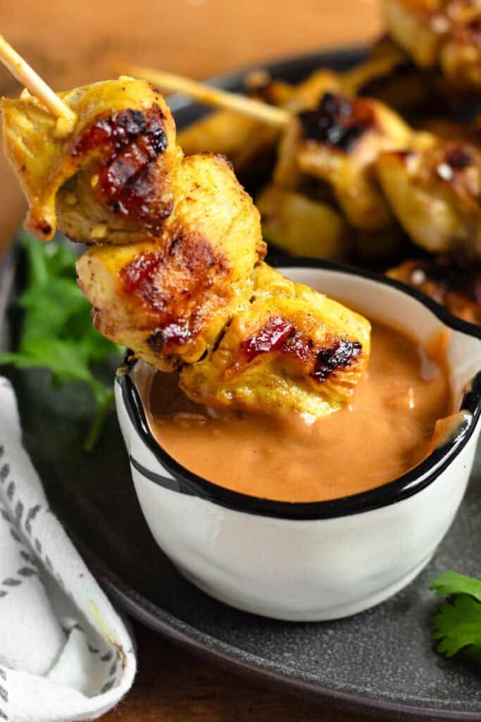 A close-up of chicken satay being dipped into a small bowl of creamy peanut sauce.
